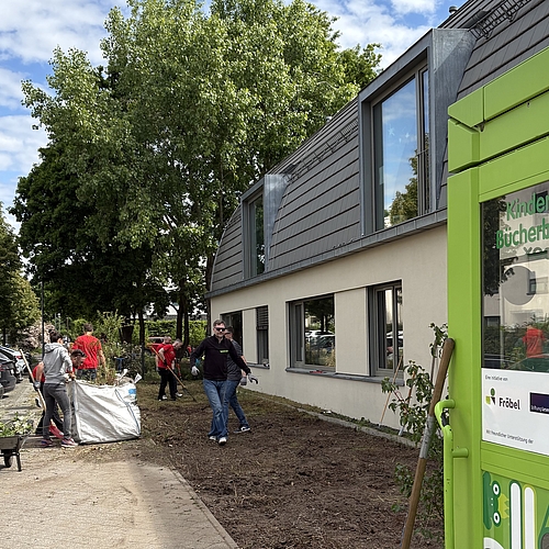 Mehrere Erwachsene arbeiten im Vorgarten des Fröbel-Kindergartens Am Volkspark. Sie sammeln Strauchschnitt und transportieren Gartenabfälle in einem großen Sack ab. Rechts im Bild steht eine grüne Kinder-Bücherbox mit der Aufschrift „Nimm meins, bring deins“. Die Aktion findet bei sonnigem Wetter direkt am Kita-Gebäude statt.