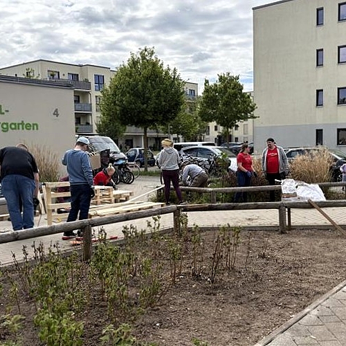 Vor dem Gebäude des Fröbel-Kindergartens Am Volkspark arbeiten zahlreiche Erwachsene an Holzkonstruktionen und im Gartenbereich. Im Vordergrund ist ein neu bepflanztes Beet zu sehen, im Hintergrund Wohnhäuser und parkende Autos. Die Szene zeigt die aktive Umgestaltung des Außenbereichs während des Audi Social Day.