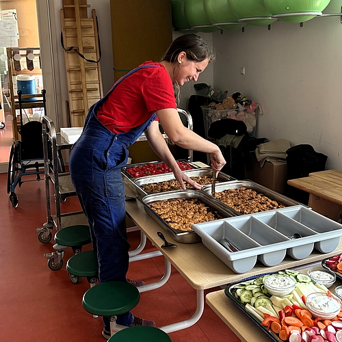 Eine Frau in rotem Shirt und blauer Latzhose richtet in einem Kita-Raum ein Buffet mit Obst, Gemüse, Dips, Kuchen und warmem Essen an. Im Vordergrund stehen Schalen mit geschnittenem Gemüse, im Hintergrund sind andere Personen und Spielmaterialien zu sehen. Die Szene zeigt die Vorbereitung der Verpflegung beim Audi Social Day.