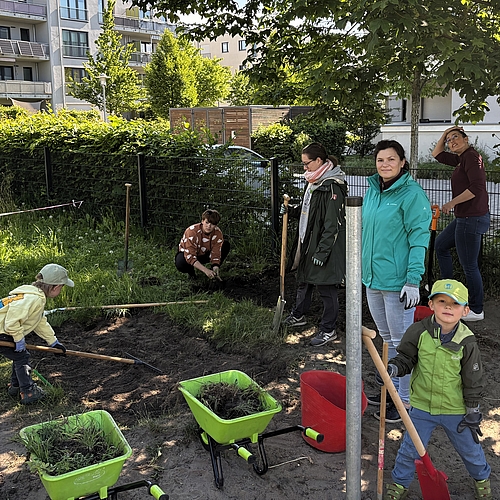 Zwei Kinder und mehrere Erwachsene arbeiten gemeinsam im Garten eines Kindergartens. Die Kinder harken Erde und befüllen kleine grüne Schubkarren mit Gras. Erwachsene unterstützen beim Graben und Vorbereiten von Beeten. Im Hintergrund Wohnhäuser, Hecken und Gartengeräte. Die Szene zeigt gemeinschaftliches Gärtnern im Schatten eines Baumes.