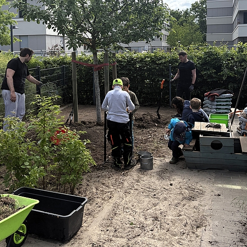 Erwachsene und Kinder arbeiten gemeinsam im hinteren Gartenteil der Kita. Ein Baum ist mit einem roten Band gesichert, Kinder spielen neben einem kleinen Holzschiff, während Erwachsene Beete vorbereiten. Schubkarren, Eimer, Blumenerde und Gartengeräte liegen bereit. Die Szene zeigt konzentrierte Gartenarbeit beim Audi Social Day.