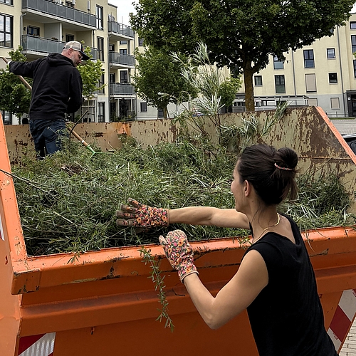Eine Frau mit bunten Gartenhandschuhen reicht abgeschnittene Zweige in einen großen Container, während ein Mann mit Rechen darin steht und das Schnittgut verteilt. Im Hintergrund sind Wohnhäuser und geparkte Autos zu sehen. Die Szene zeigt gemeinschaftliches Arbeiten beim Audi Social Day am Fröbel-Kindergarten Am Volkspark.