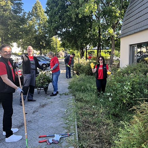 Mehrere Erwachsene in roten Audi-T-Shirts arbeiten im Vorgarten des Fröbel-Kindergartens Am Volkspark. Sie schneiden Büsche, harken Laub und schieben eine Schubkarre mit Grünschnitt. Im Hintergrund sind parkende Autos und Bäume zu sehen. Alle wirken fröhlich und motiviert.