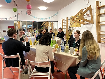 Businessfrühstück im Fröbel-Kindergarten Am Volkspark. Im Bewegungsraum des Kindergartens sitzen Menschen an gedeckten Tischen, frühstücken und unterhalten sich
