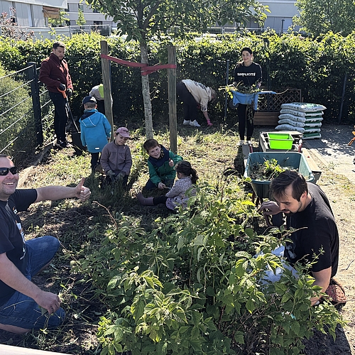 Erwachsene und Kinder arbeiten gemeinsam im Garten eines Kindergartens. Einige Kinder sitzen lachend im Gras, während andere zusammen mit Erwachsenen Unkraut jäten, Sträucher schneiden und Schubkarren befüllen. Im Hintergrund stehen Gartenmaterialien wie Säcke mit Erde und Werkzeuge bereit. Ein Mann im Vordergrund zeigt lächelnd den Daumen hoch – die Stimmung ist fröhlich und tatkräftig.