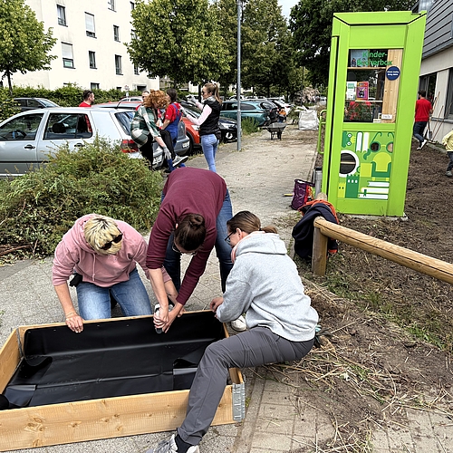 Drei Erwachsene befestigen eine schwarze Folie in einem Holzrahmen für ein Hochbeet. Im Hintergrund stehen Eltern an parkenden Autos, Kinder spielen auf der freien Fläche vor der Kita. Rechts im Bild eine grüne Kinder-Bücherbox und das frisch gerodete Beet entlang des Gebäudes.