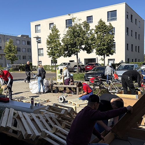 Erwachsene bauen auf einem Parkplatz Holzmöbel aus Paletten, sägen, schrauben und schleifen. Kinder fahren im Hintergrund Fahrrad. Im Vordergrund liegt gestapeltes Holz. Die Szene zeigt den Möbelbau beim Audi Social Day am Fröbel-Kindergarten Am Volkspark.
